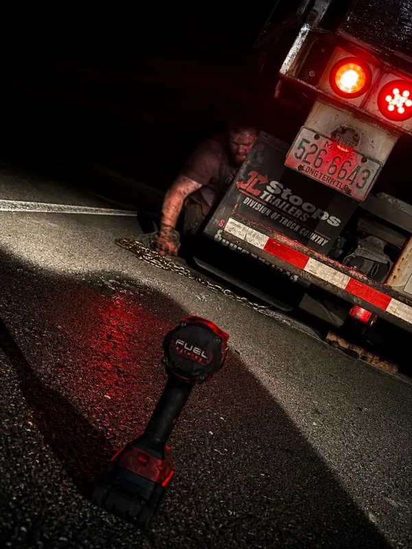 Mechanic working under the rear of a flatbed trailer at night, impact wrench on the asphalt